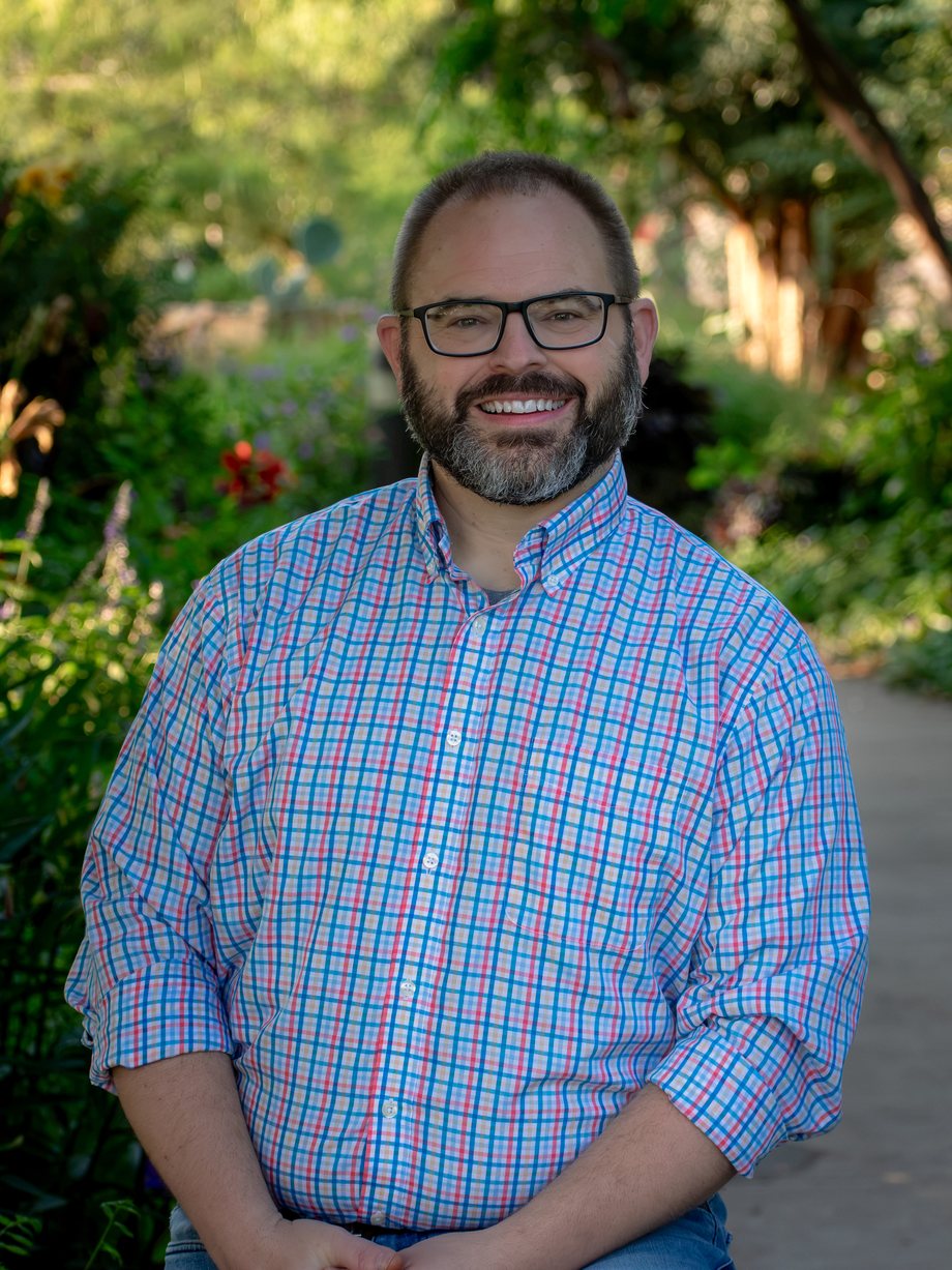 Blake Grisham, smiling, in glasses and a blue and red checked shirt, on a garden path lined with summer flowers.