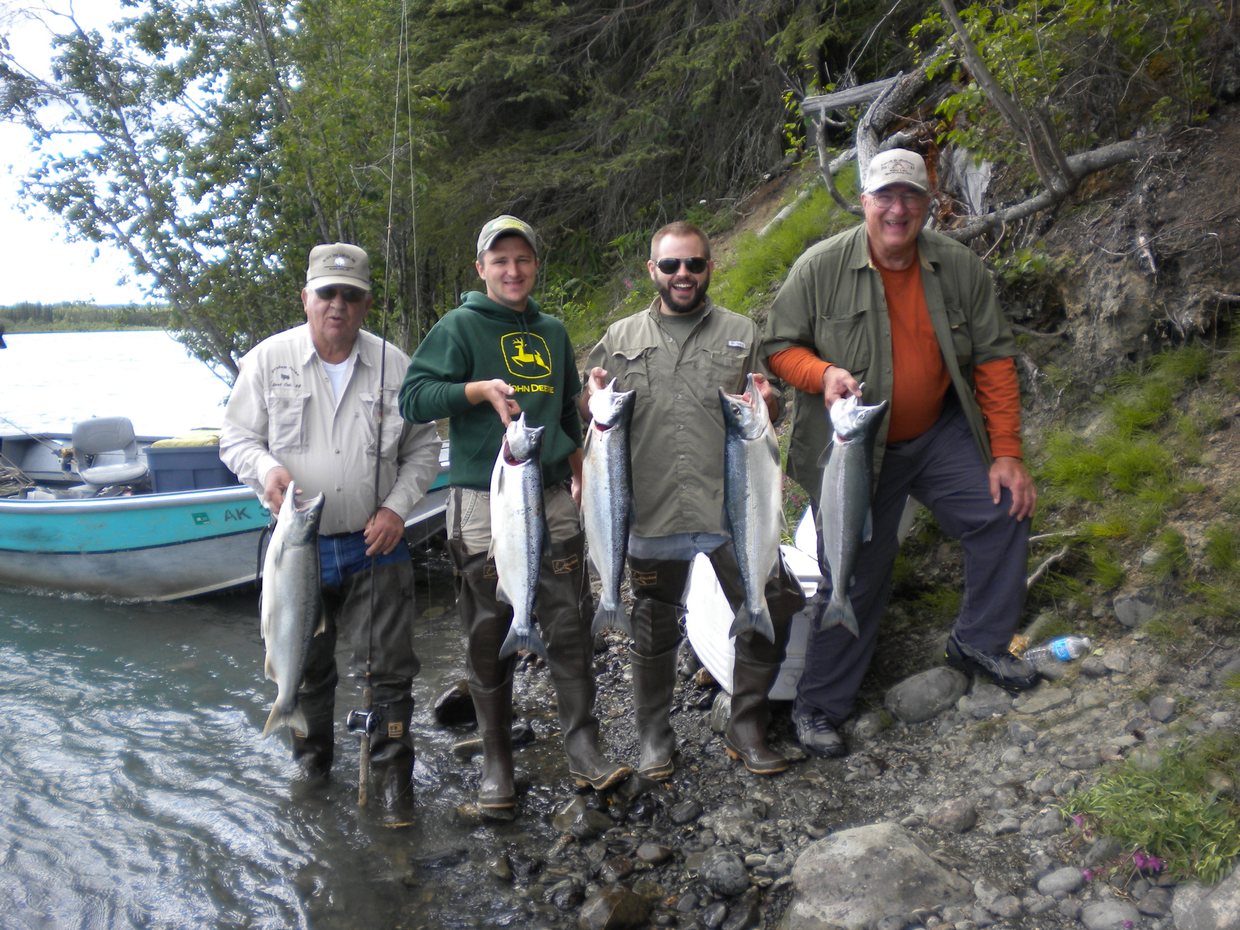 Four men stand on a rocky riverbank, each holding a silver salmon, with a drift boat pulled up behind them and a wooded slope above. From left: Daddy Bob, Blaine, Blake, and Uncle Lynn.