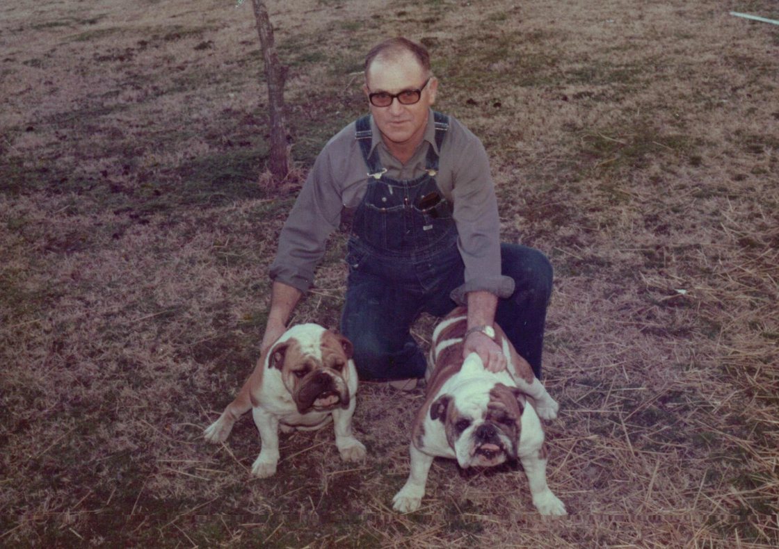 Pa Paw kneels in a dry pasture in glasses, a work shirt, and overalls, a hand on each of his two English bulldogs.