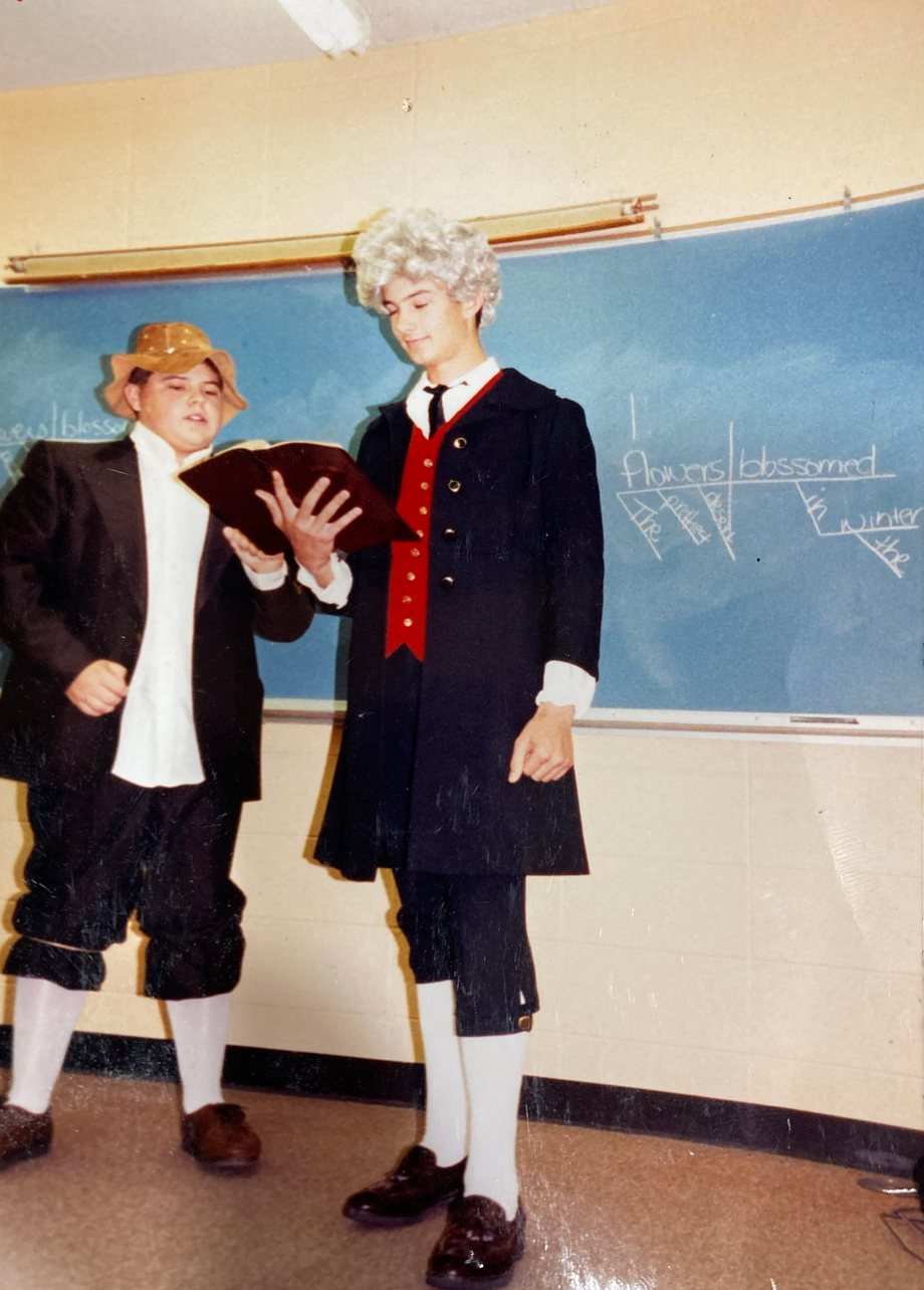 Two teenage boys in colonial costume stand at the front of a classroom, reading from one open book. The boy on the left wears a gold hat, a black jacket, and black breeches gathered at the knee with a rubber band; the taller boy on the right wears a white curled wig, a long dark coat over a red vest, and knee breeches. Behind them, a sentence is diagrammed on the chalkboard.