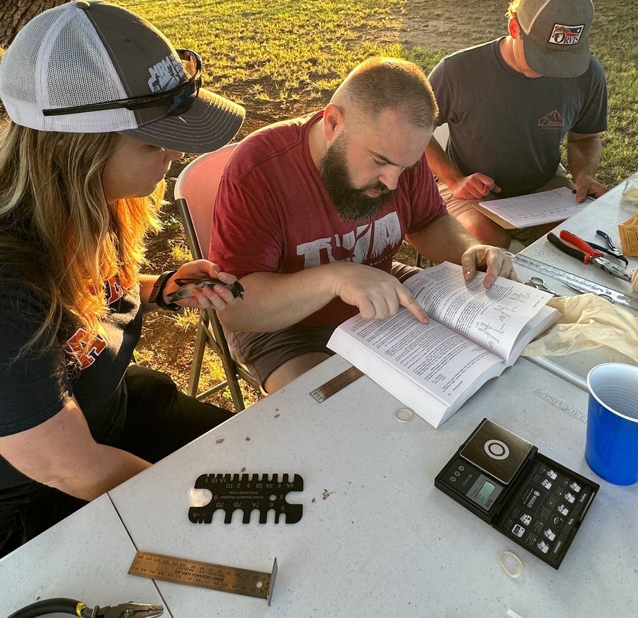 Blake Grisham reads a banding guide at a table at sunrise while one colleague holds a Cliff Swallow and another records data, with banding tools laid out.