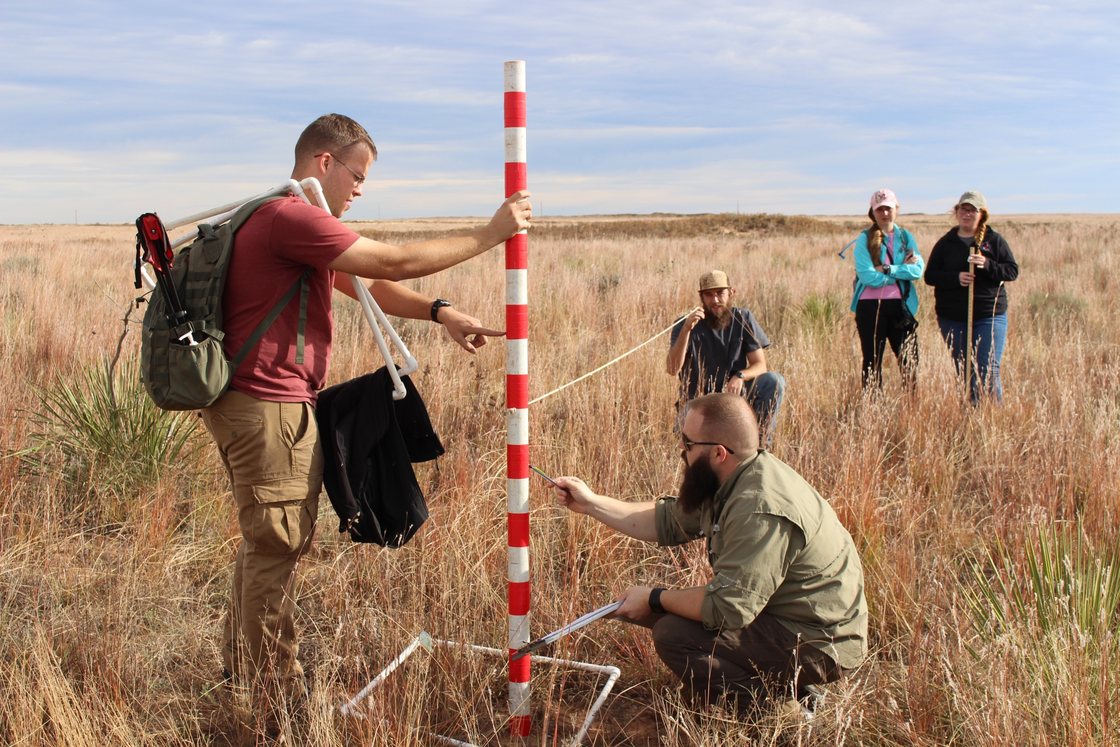 Blake Grisham kneels with a data sheet at a vegetation sampling frame while a student holds a striped measuring pole; more students work in the field behind.