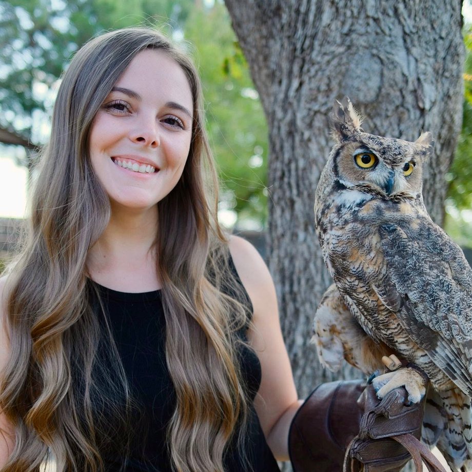 Sarah Hollers smiles beside a tree, a Great Horned Owl perched on the leather glove on her left hand.